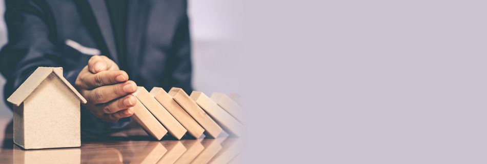 A person is blocking the dominoes that fall one after another toward a house model.