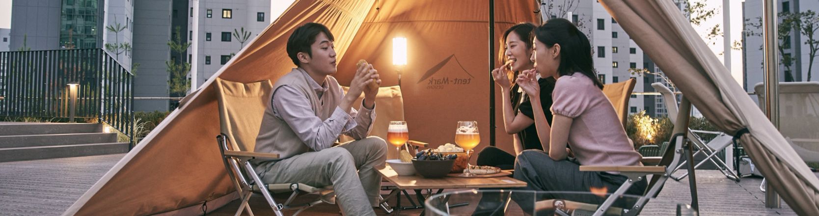 세 사람이 텐트 앞 테이블과 의자에 둘러앉아 음식을 나누어 먹고 있습니다.